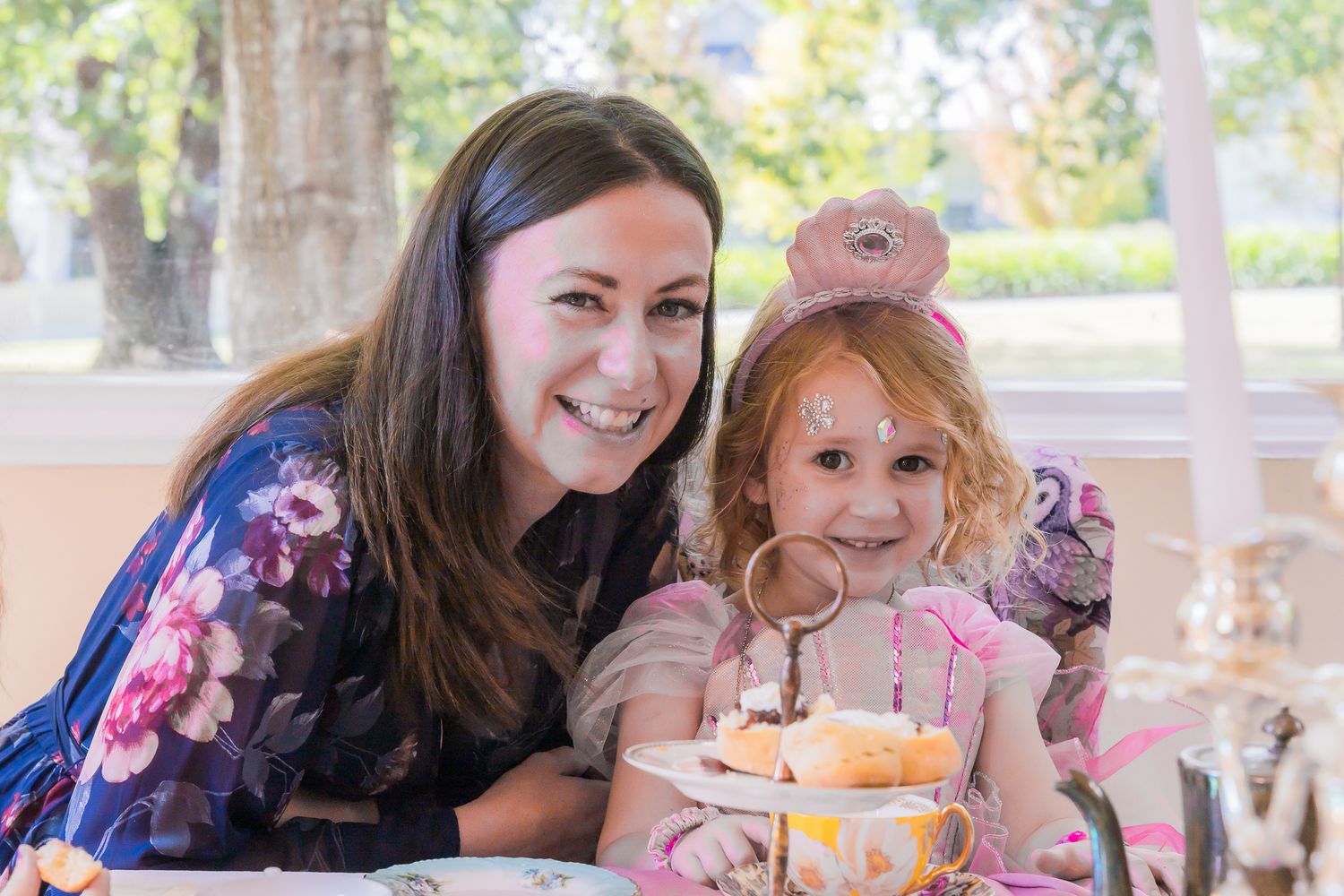 Smiling mother and young daughter enjoying afternoon tea party with cupcakes together.