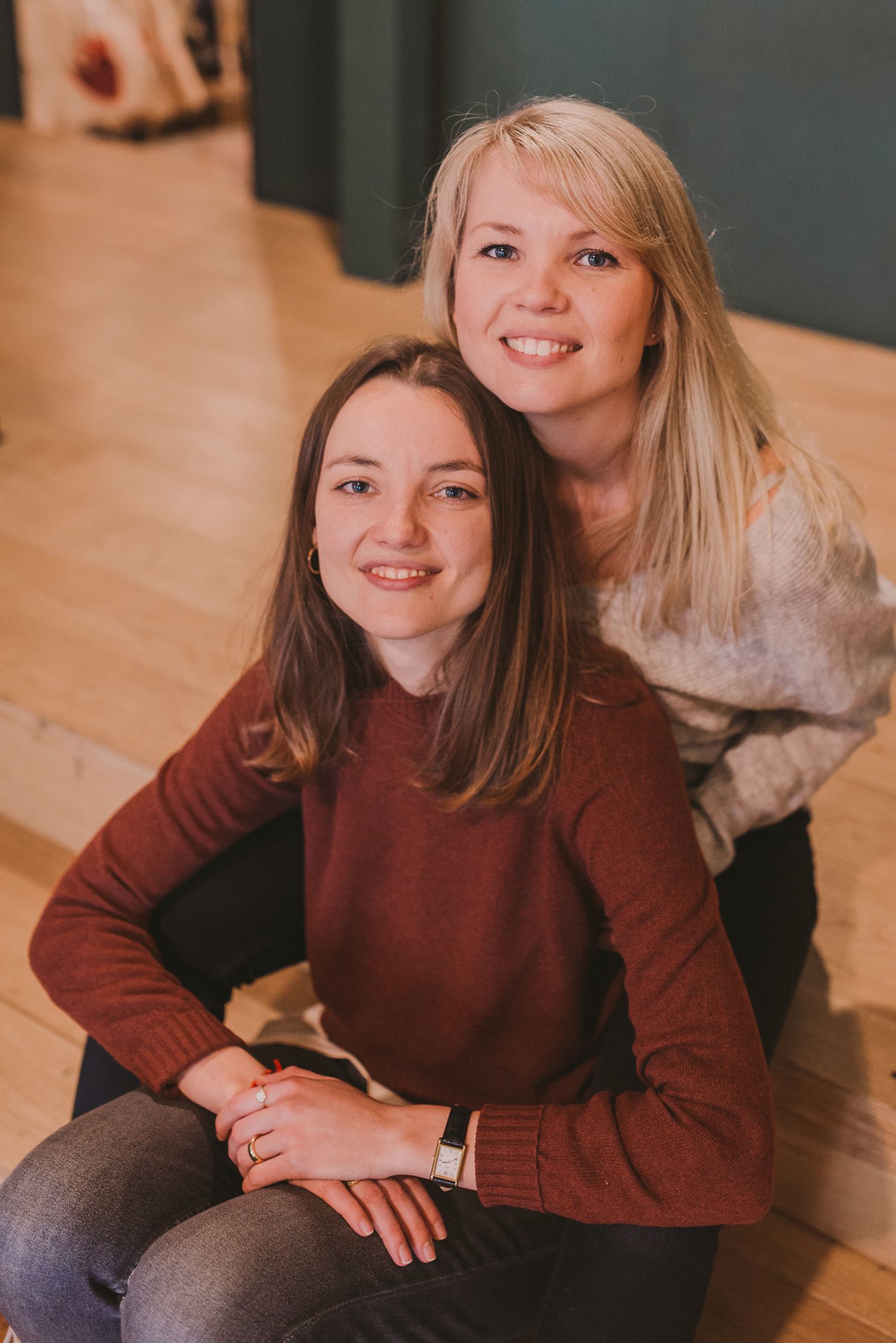 Two friends in burgundy and gray sweaters pose together on wooden flooring.