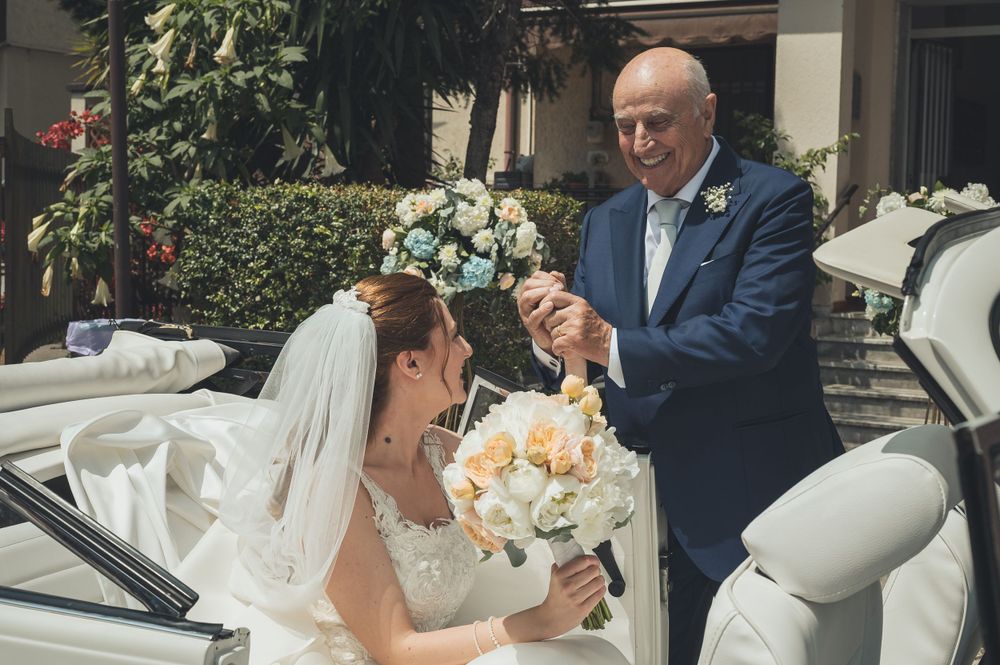 Father helping bride out of white vintage car holding her bouquet of pastel flowers.