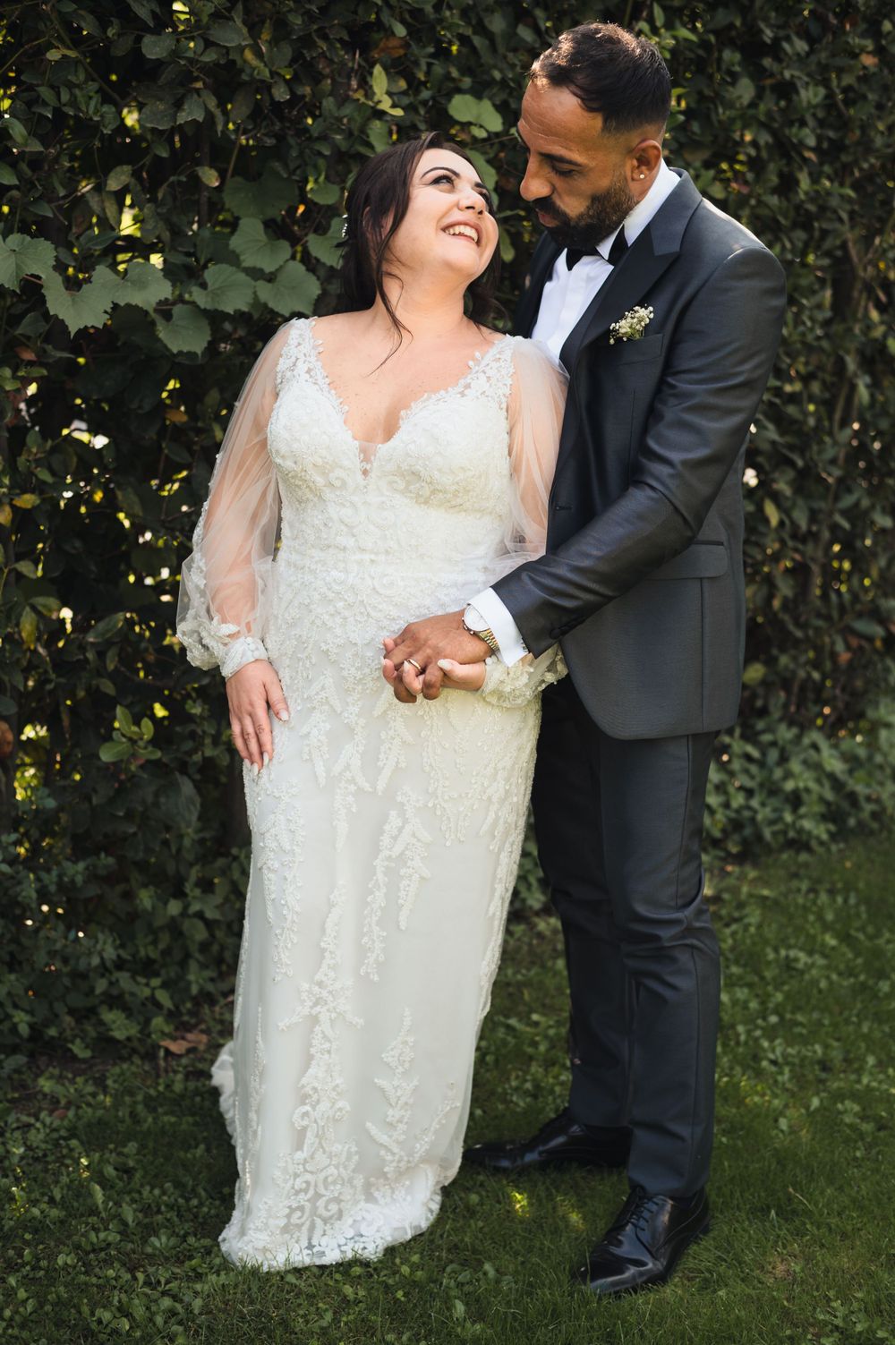 Romantic wedding couple embracing outdoors against green hedge background.