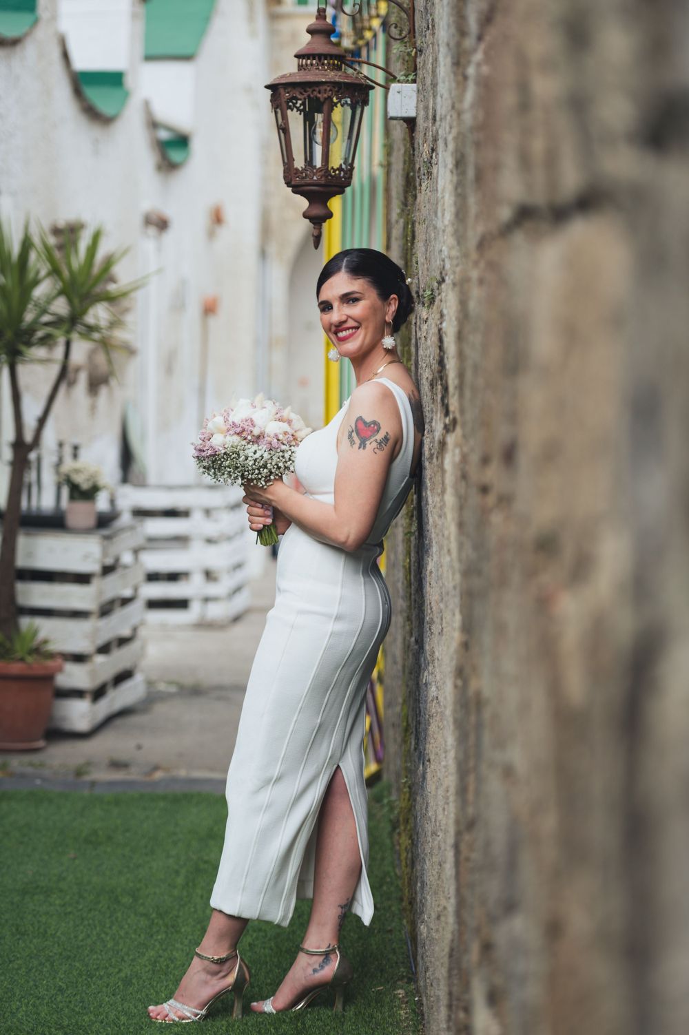 A person in a white dress poses with flowers against a wall with vintage lantern fixture.
