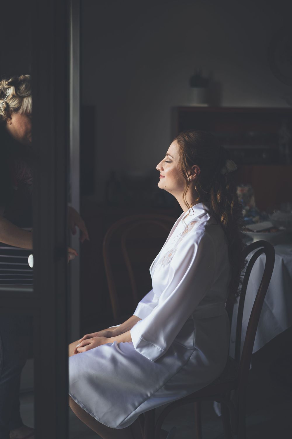 Bride in white robe sitting in chair getting ready with natural light streaming in.