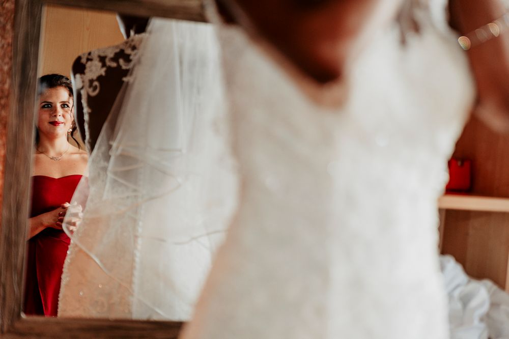 An elegant wedding dress is reflected in a vintage mirror with a person in red visible in the background.