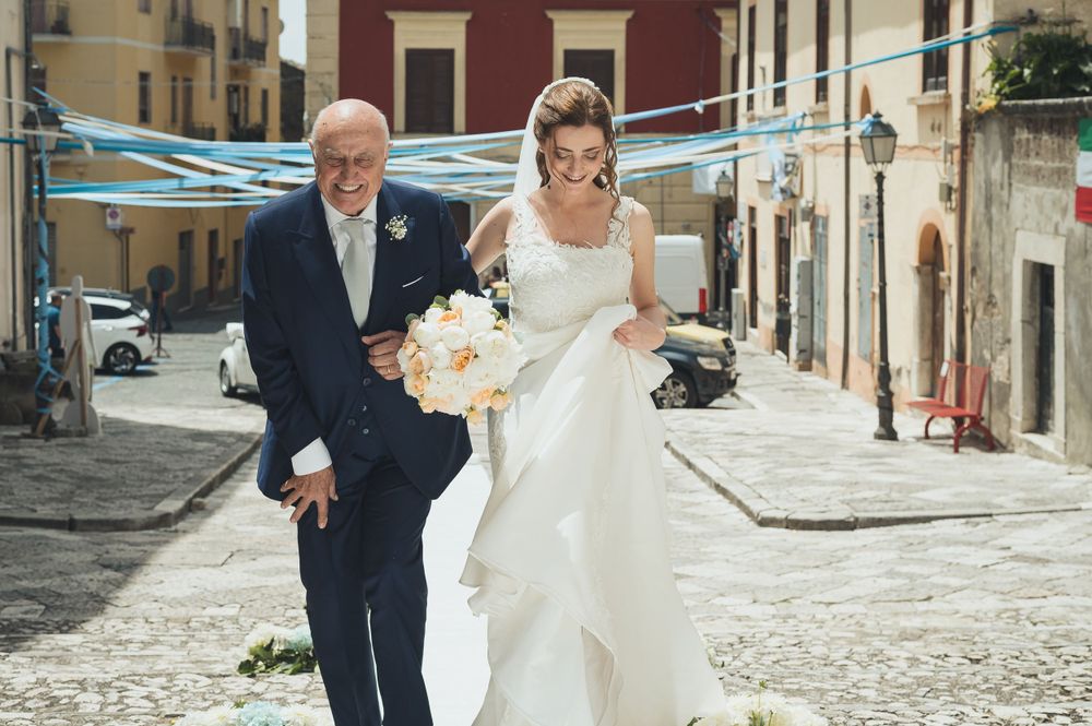 Bride in white gown walking with father through charming Italian village street.