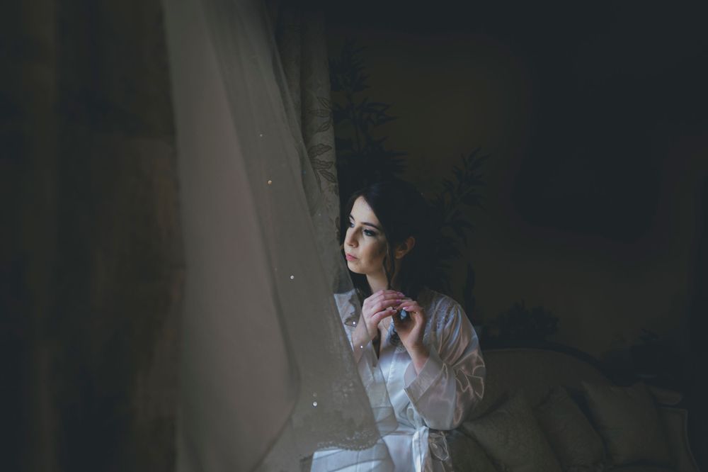 Elegant bride in white robe gazing thoughtfully out a window in soft moody light.