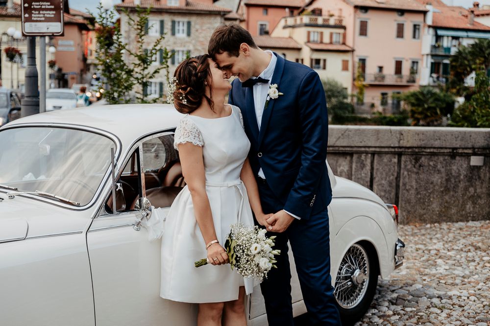 A newlywed couple poses romantically next to a vintage white car in a European setting.