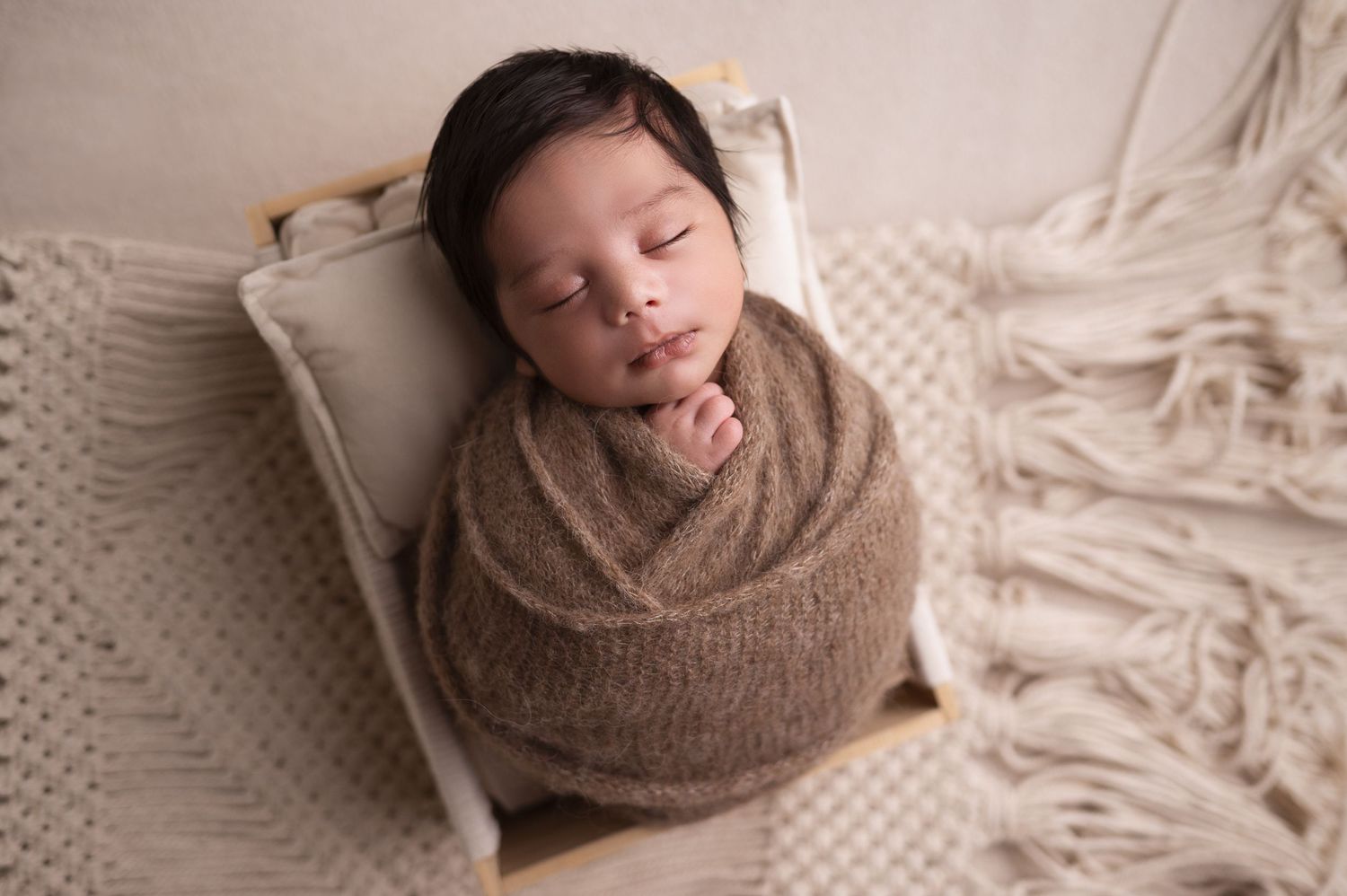 Artistic newborn photography with baby wrapped in brown swaddle against cream background.