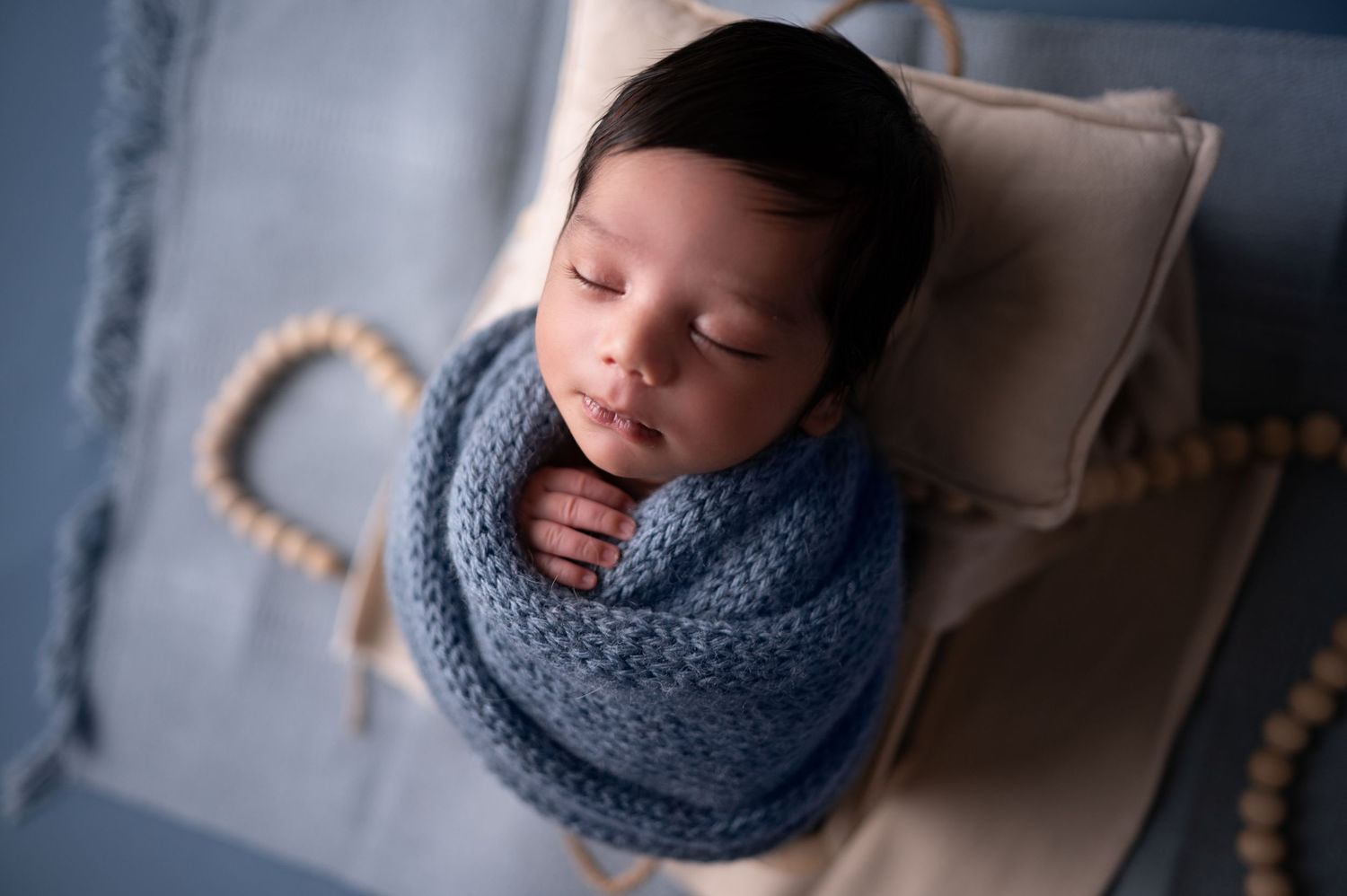 Newborn wrapped in blue knit blanket sleeping on neutral pillow.