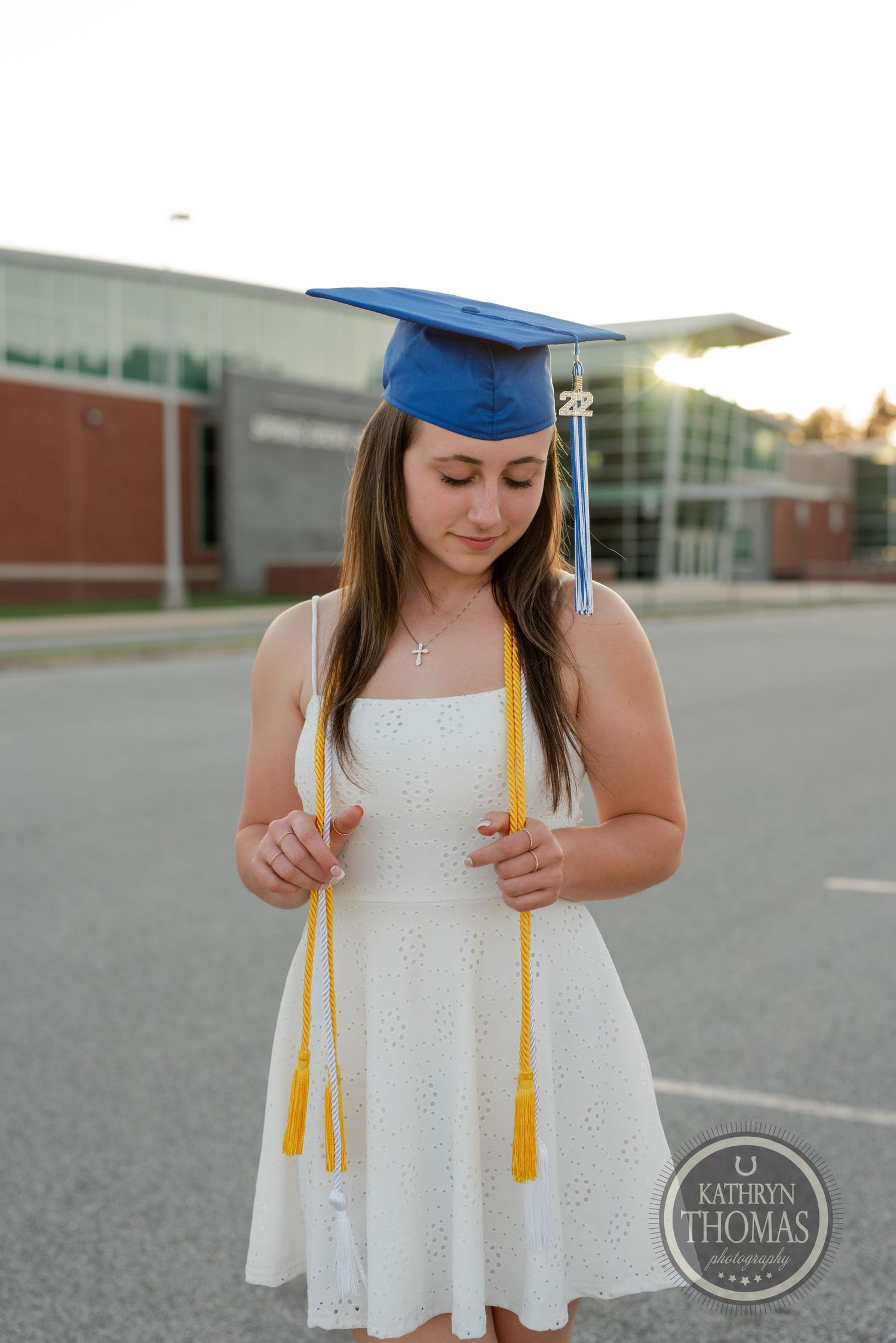 Graduate in white eyelet dress holds gold tassel while wearing blue cap and gown.