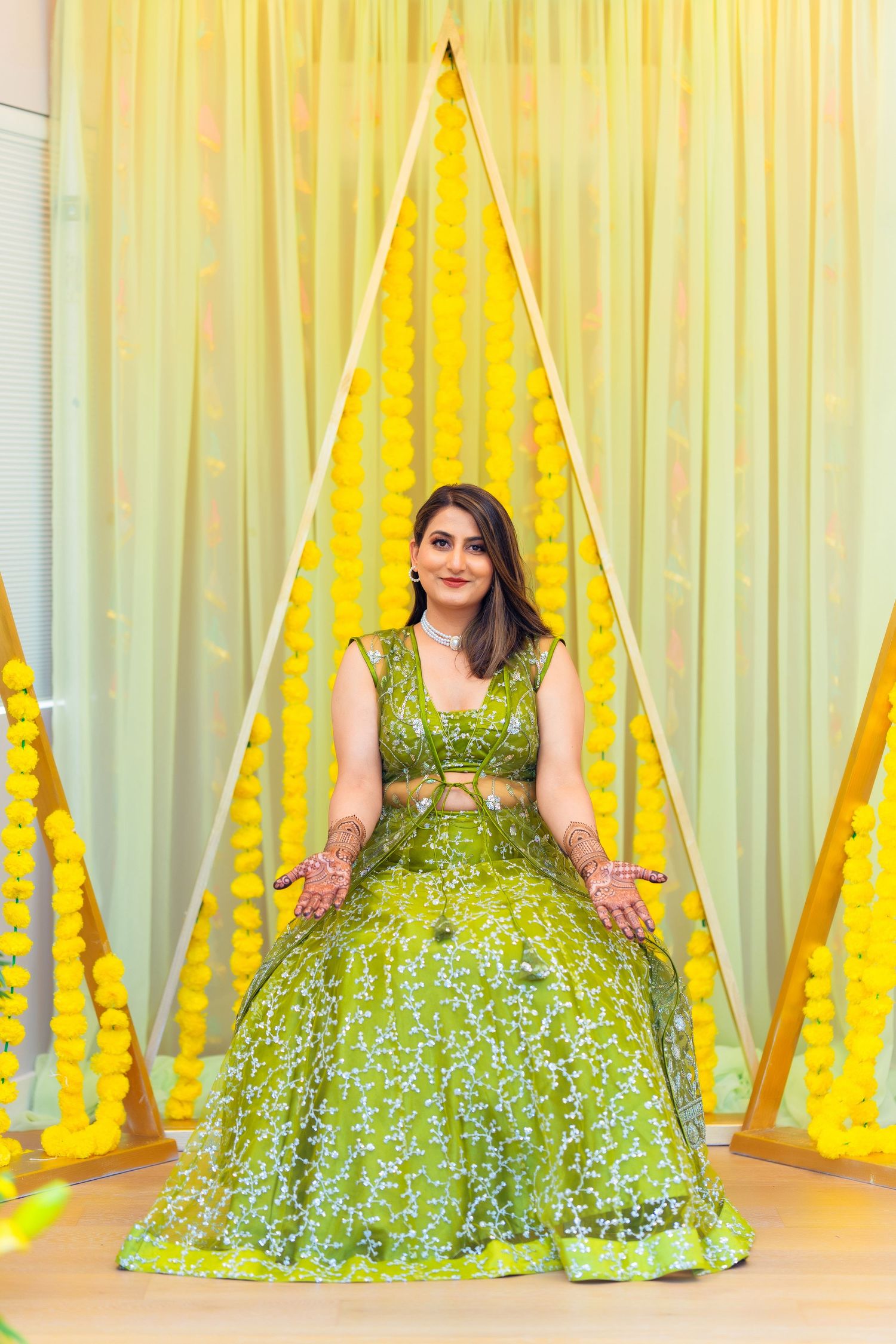 Traditional bridal pose in sparkling green lehenga against yellow and white draped backdrop.