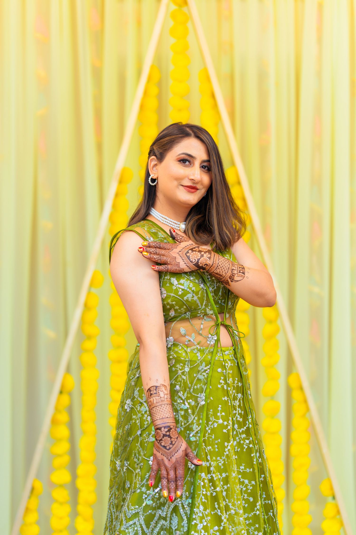 Woman in sparkly green lehenga poses with henna-adorned hands against yellow curtains.
