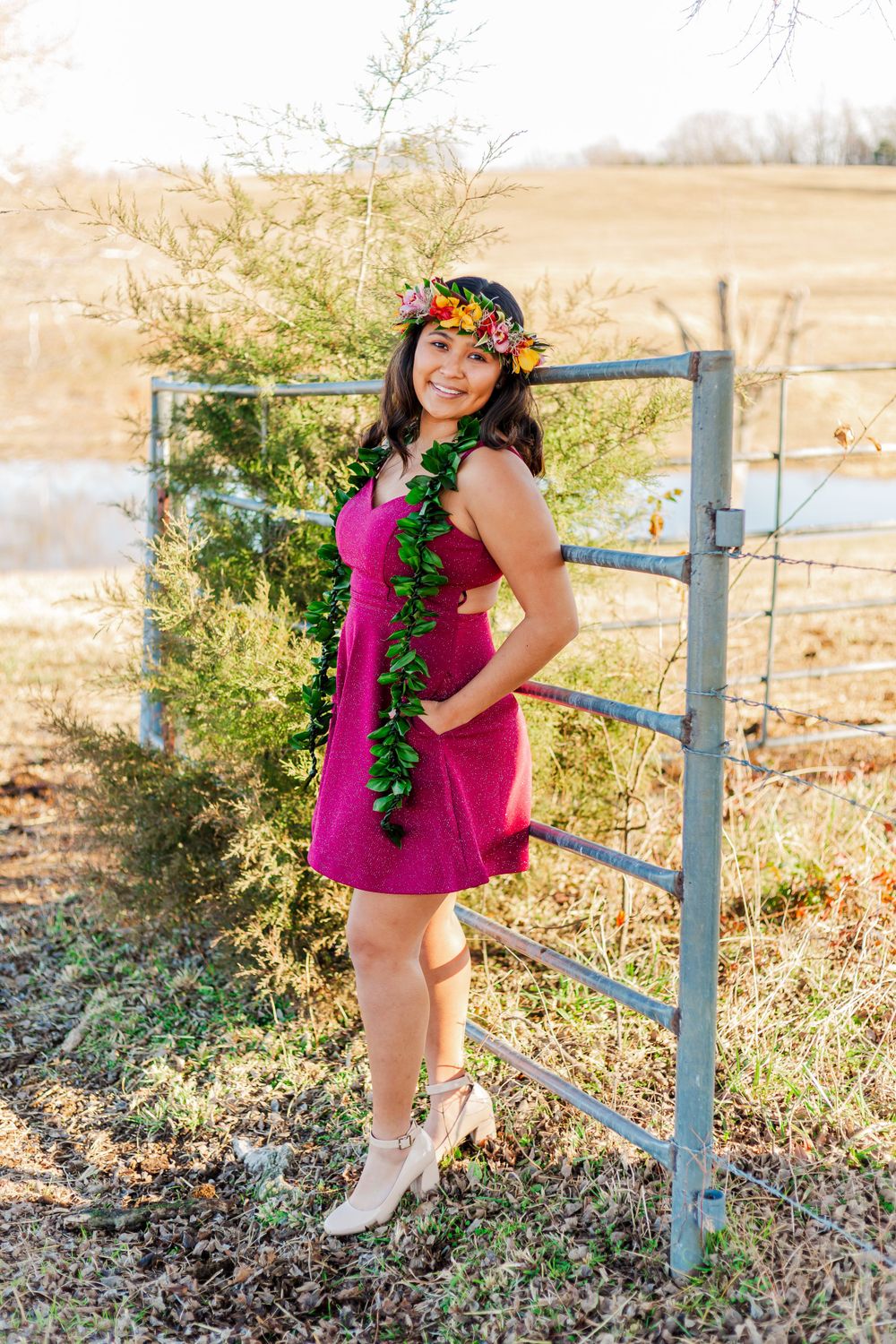 Figure in magenta dress poses by rustic fence wearing floral crown and green lei.