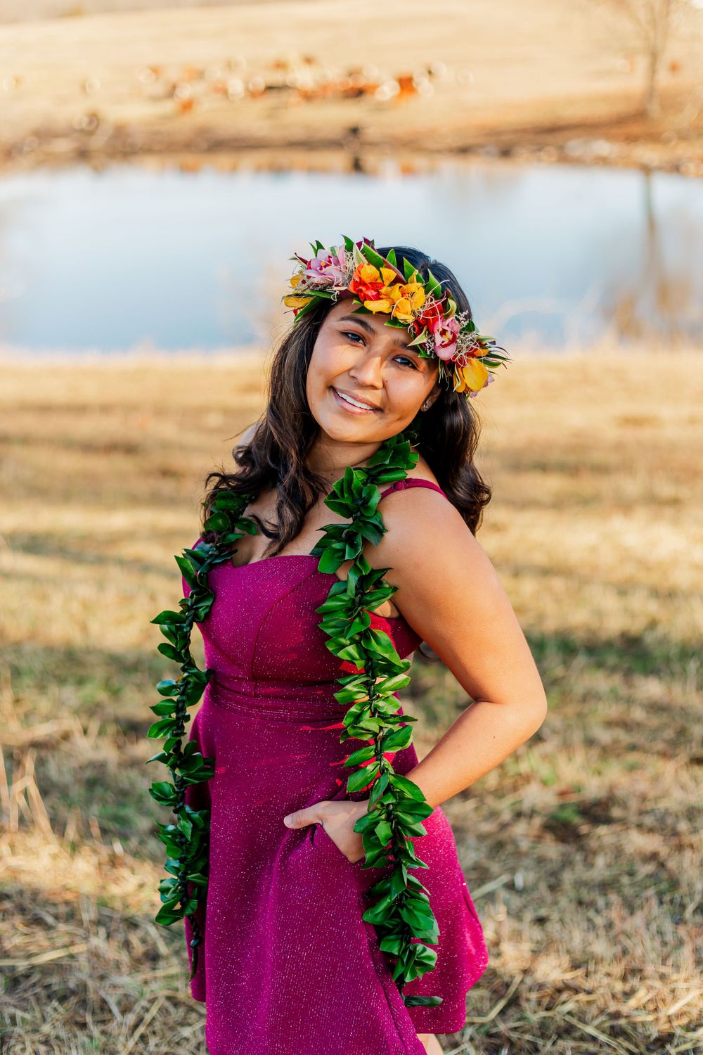 Portrait in magenta dress with flower crown and green lei against scenic countryside backdrop.