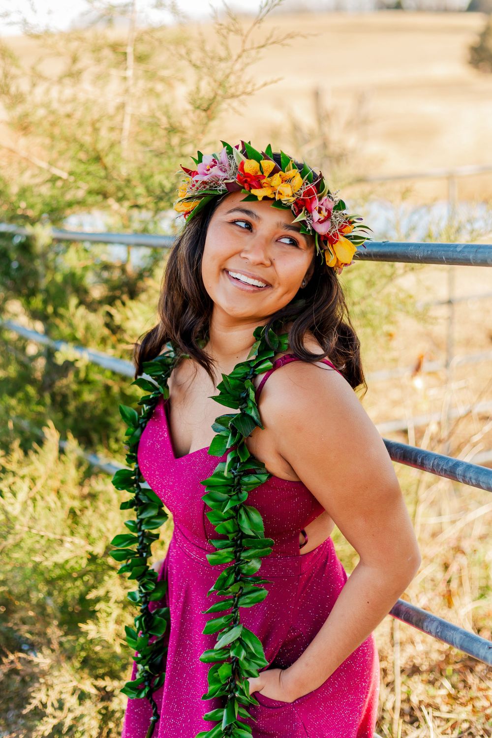 Figure wearing tropical flower crown and green lei poses in magenta dress against natural setting.