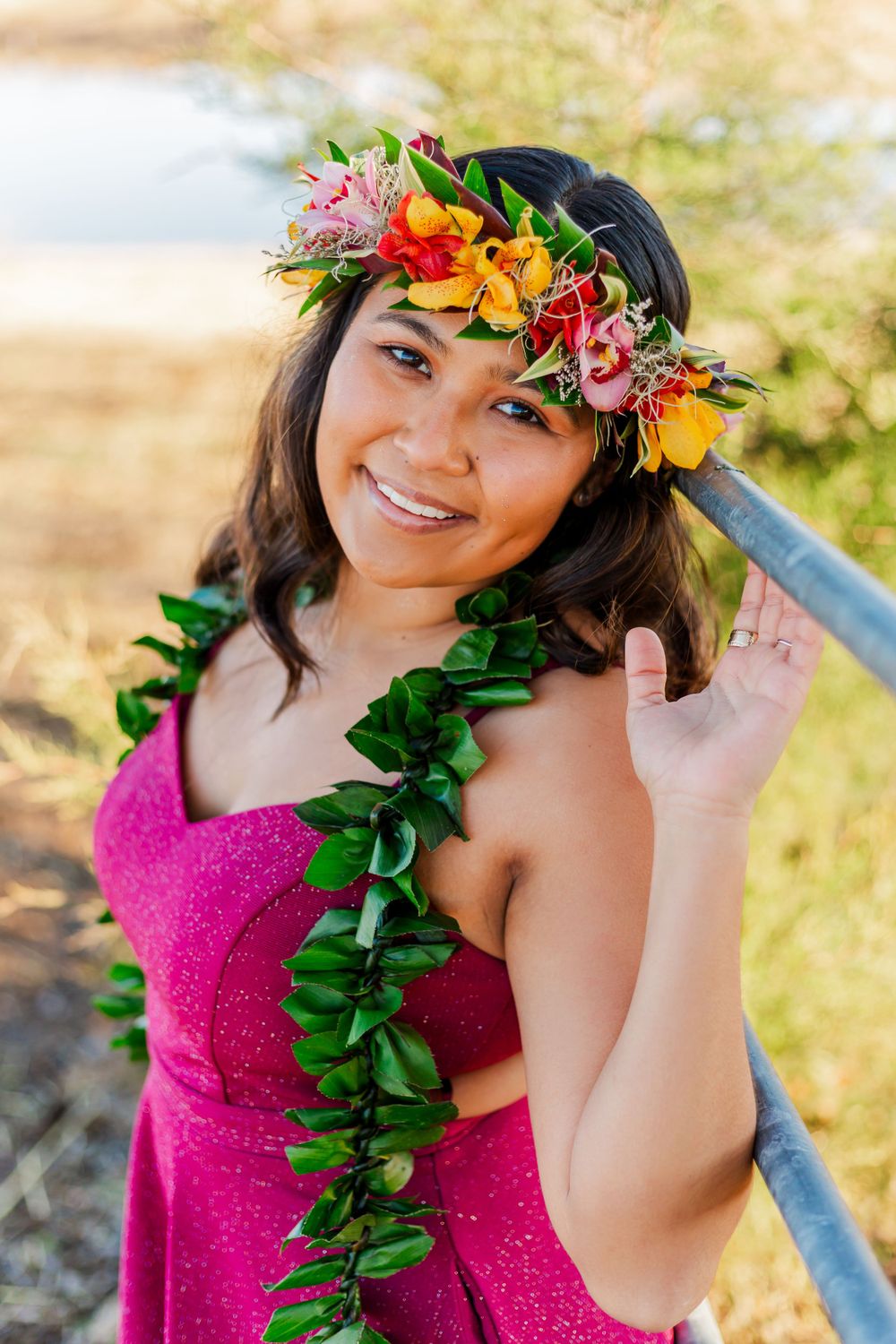 Individual in a hot pink dress adorned with tropical floral crown and green lei poses outdoors.