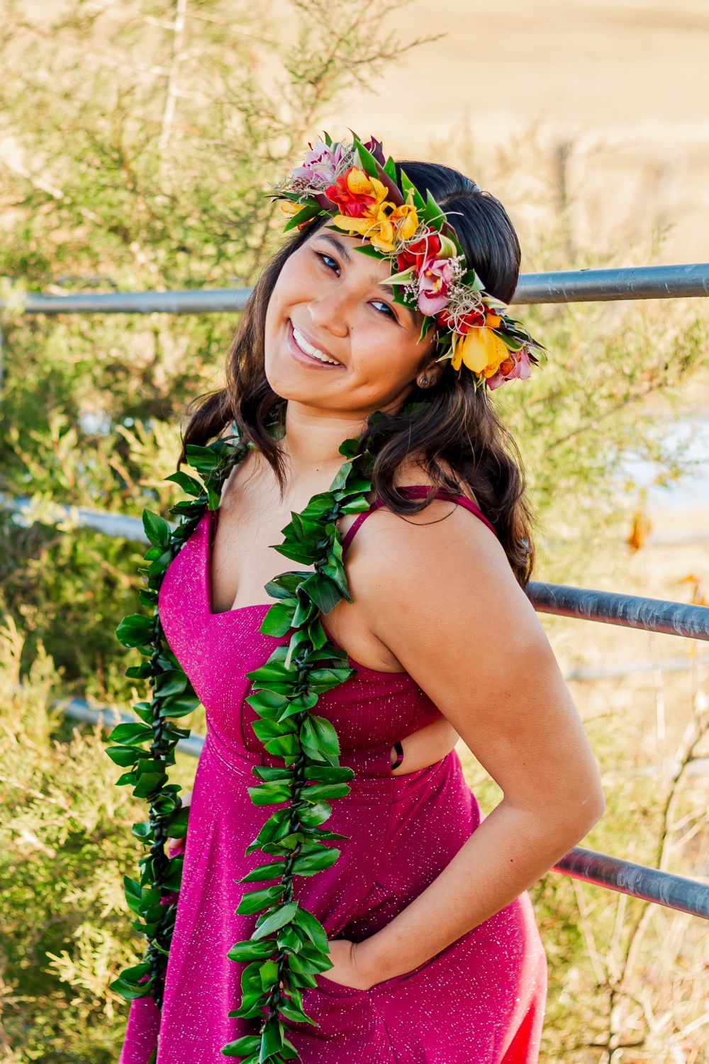Person in bright pink dress with tropical lei and floral headpiece poses against rural backdrop.