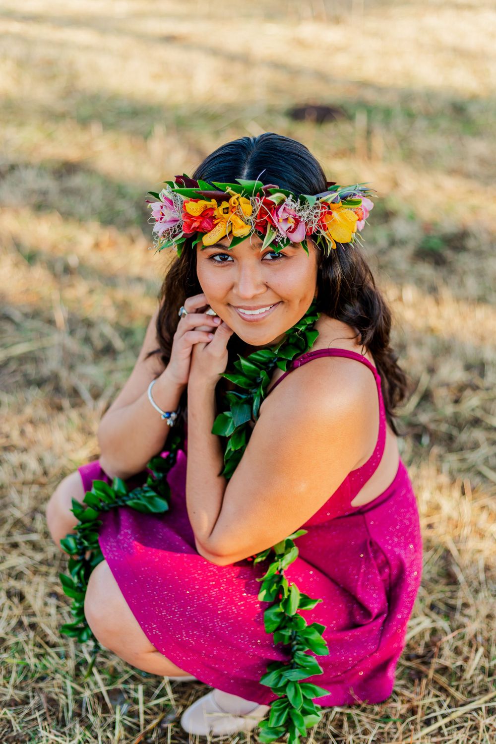 Candid portrait wearing flower crown and magenta dress while crouching in sunny field.