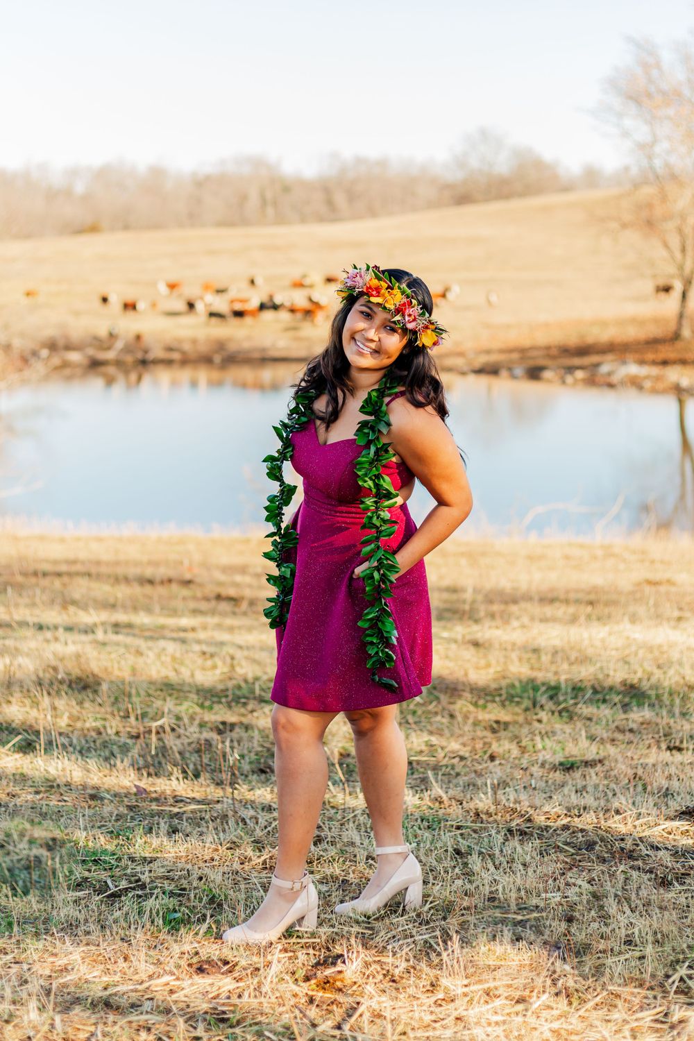 Woman in magenta dress and tropical accessories poses by pond in rural setting.