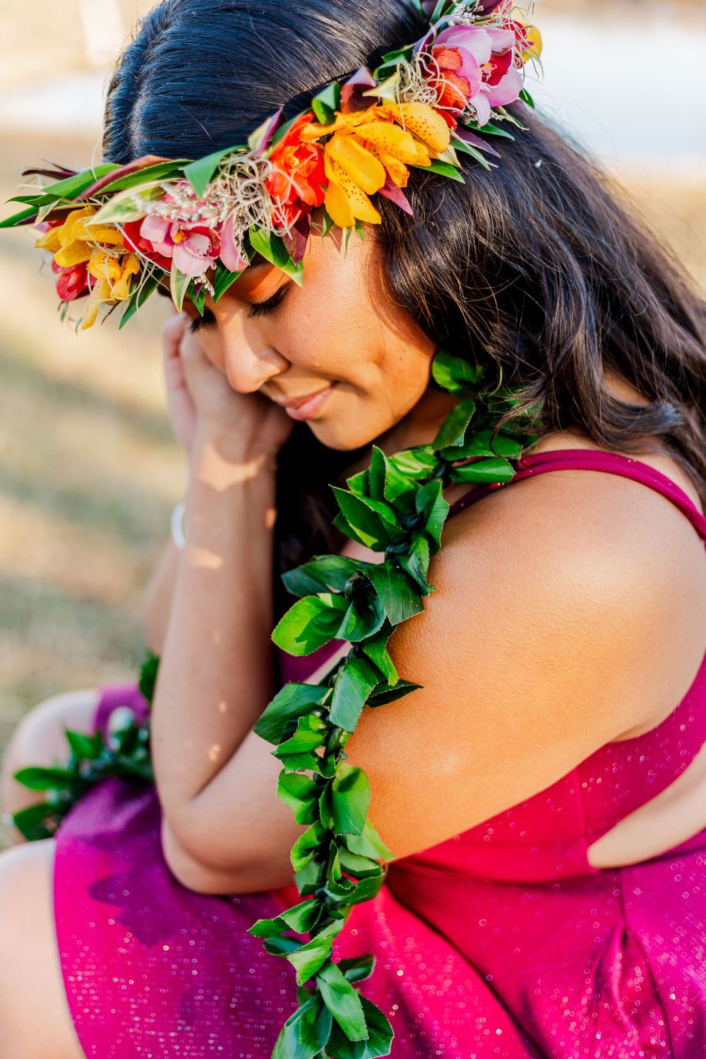 Person wearing a vibrant floral crown and green lei poses thoughtfully in a magenta dress.