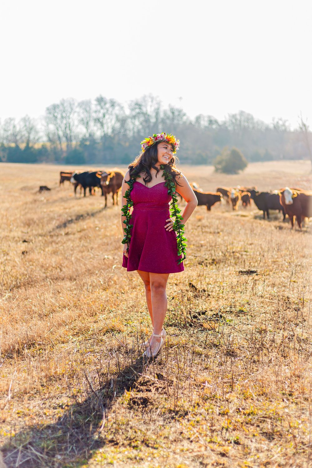 Woman in magenta dress and floral crown standing in golden field with cattle grazing in background.