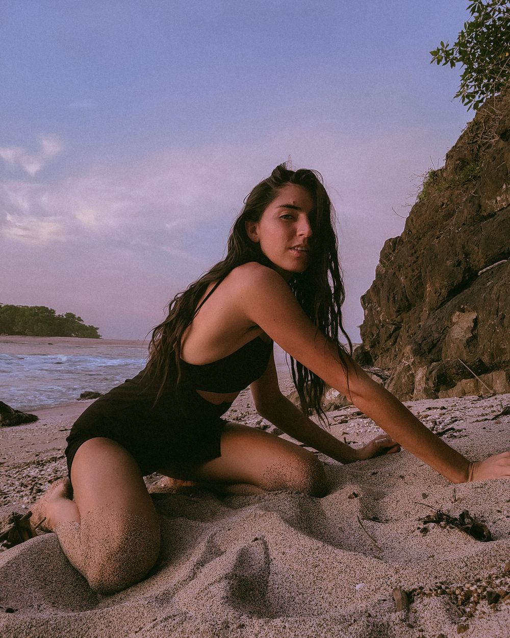 Figure poses on sandy beach with rocky cliff backdrop during purple sunset.