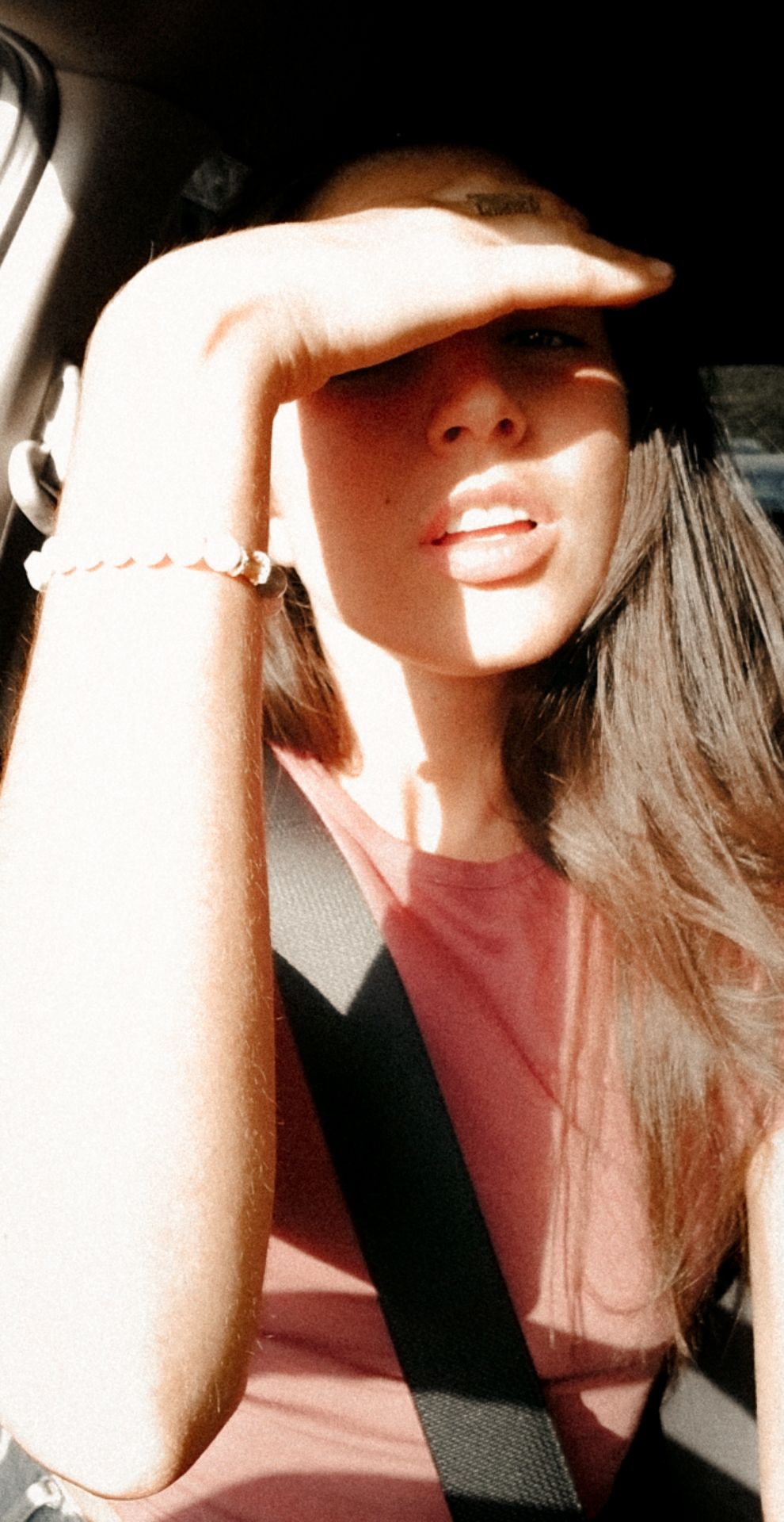 Sun-drenched car selfie with hand shielding eyes from bright natural light.