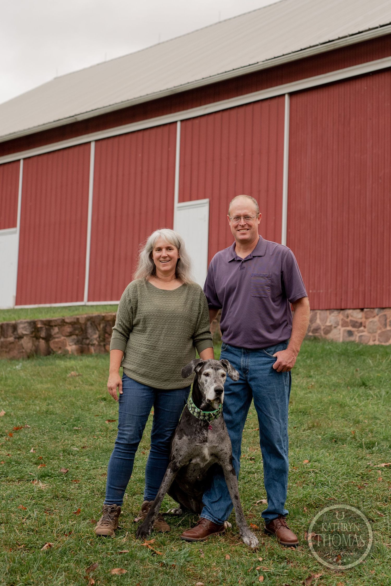 A couple poses with their black dog in front of a traditional red barn with stone foundation.