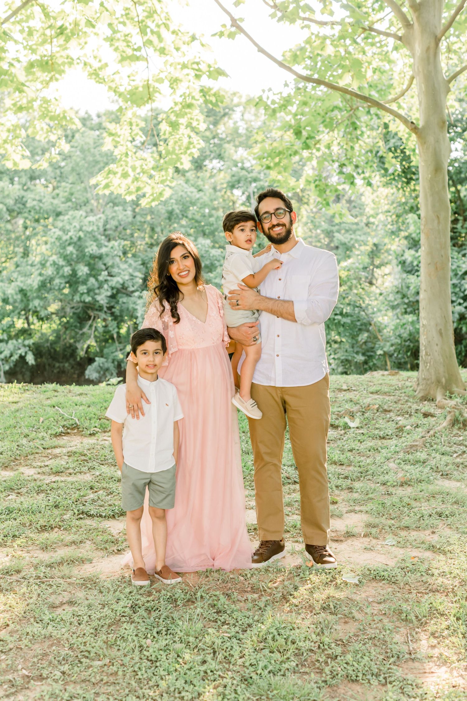 A family of four poses together in a sunlit outdoor setting with trees in background.