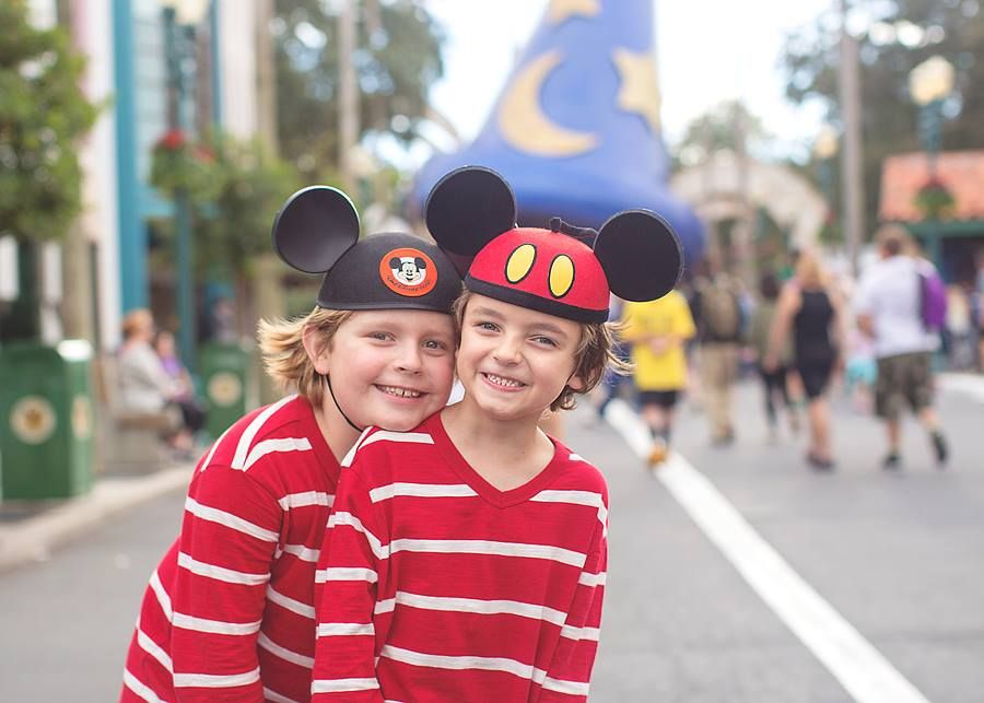 Disney park visitors wearing striped shirts and Mickey ear hats smile on Hollywood Studios street.