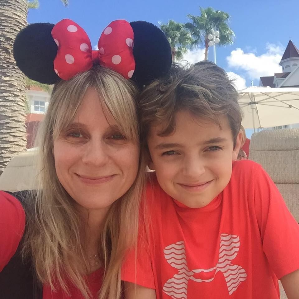 Visitors wearing Minnie Mouse ears pose for photo at Disney theme park with palm trees in background.