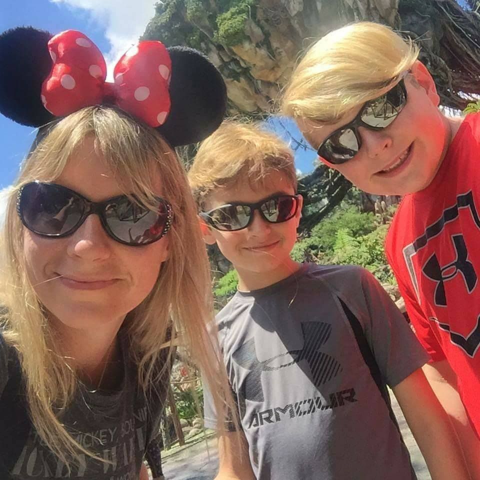Disney theme park visitors wearing Minnie ears and sunglasses pose for selfie with tropical scenery.