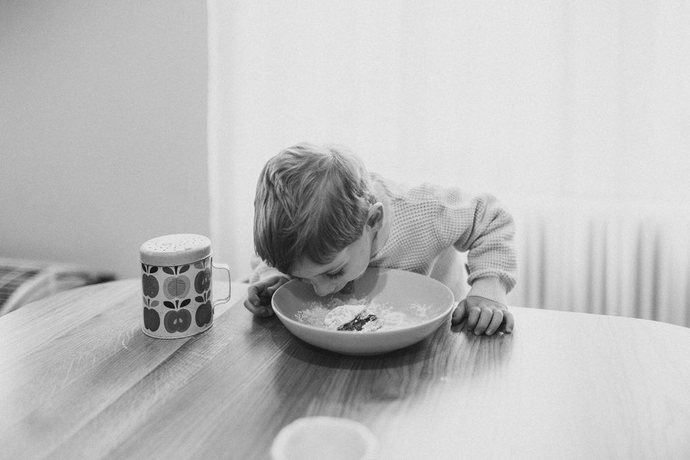 Black and white image of a child eating from a plate at a wooden table with a can nearby.