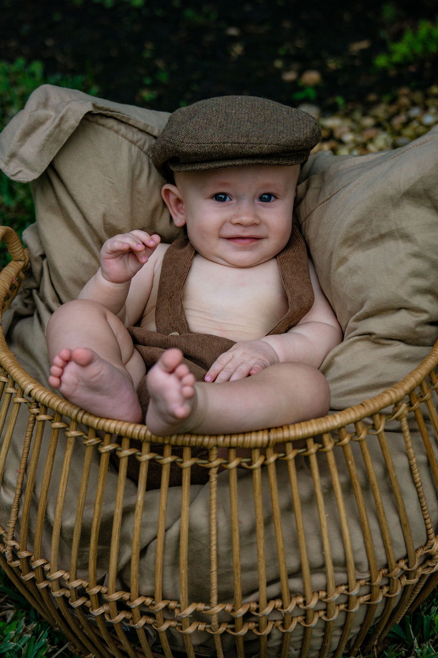 Baby wearing newsboy cap and suspenders sitting in wicker chair outdoors.