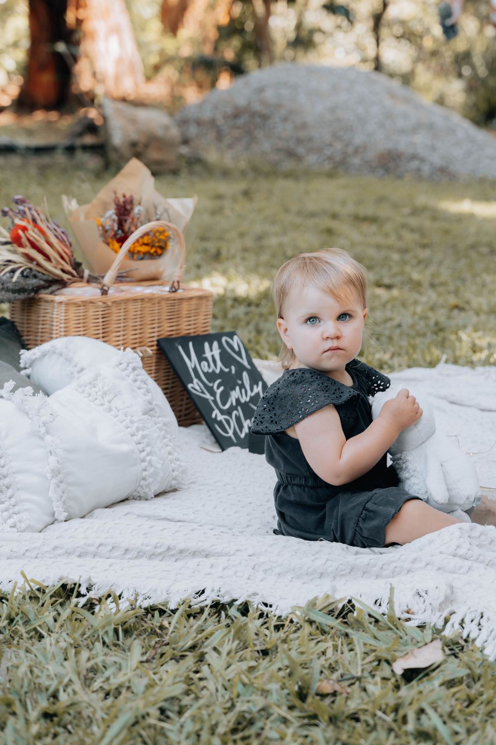 A toddler in black sitting on a white picnic blanket next to a wicker basket.