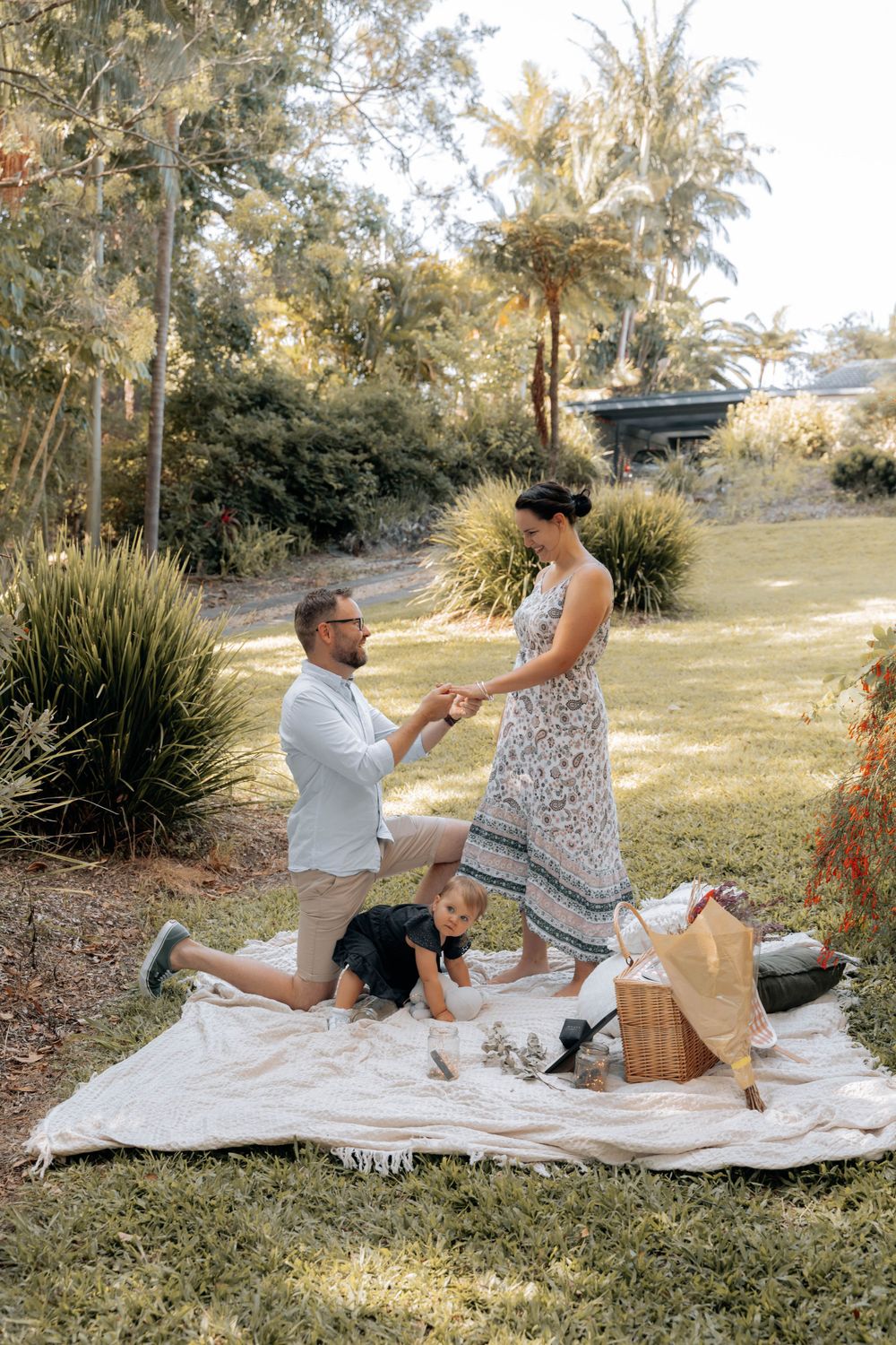 A romantic proposal scene captured in a garden setting with a picnic blanket and wicker basket.