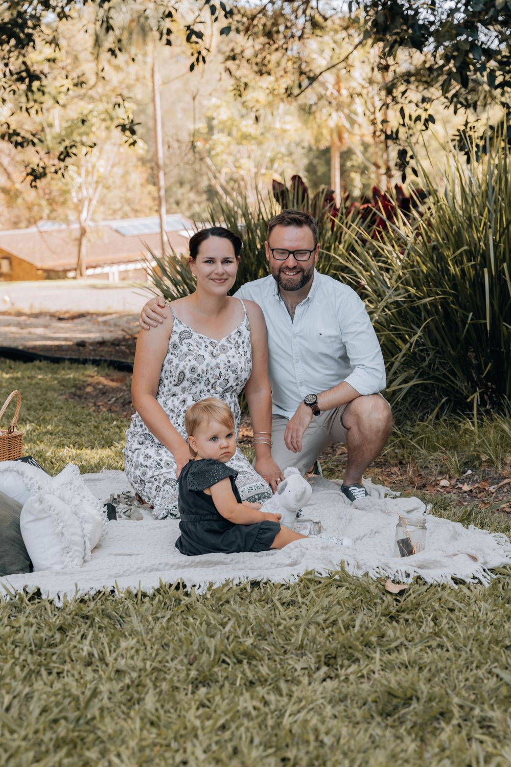 A family picnic portrait on a white blanket in a sunny garden setting.