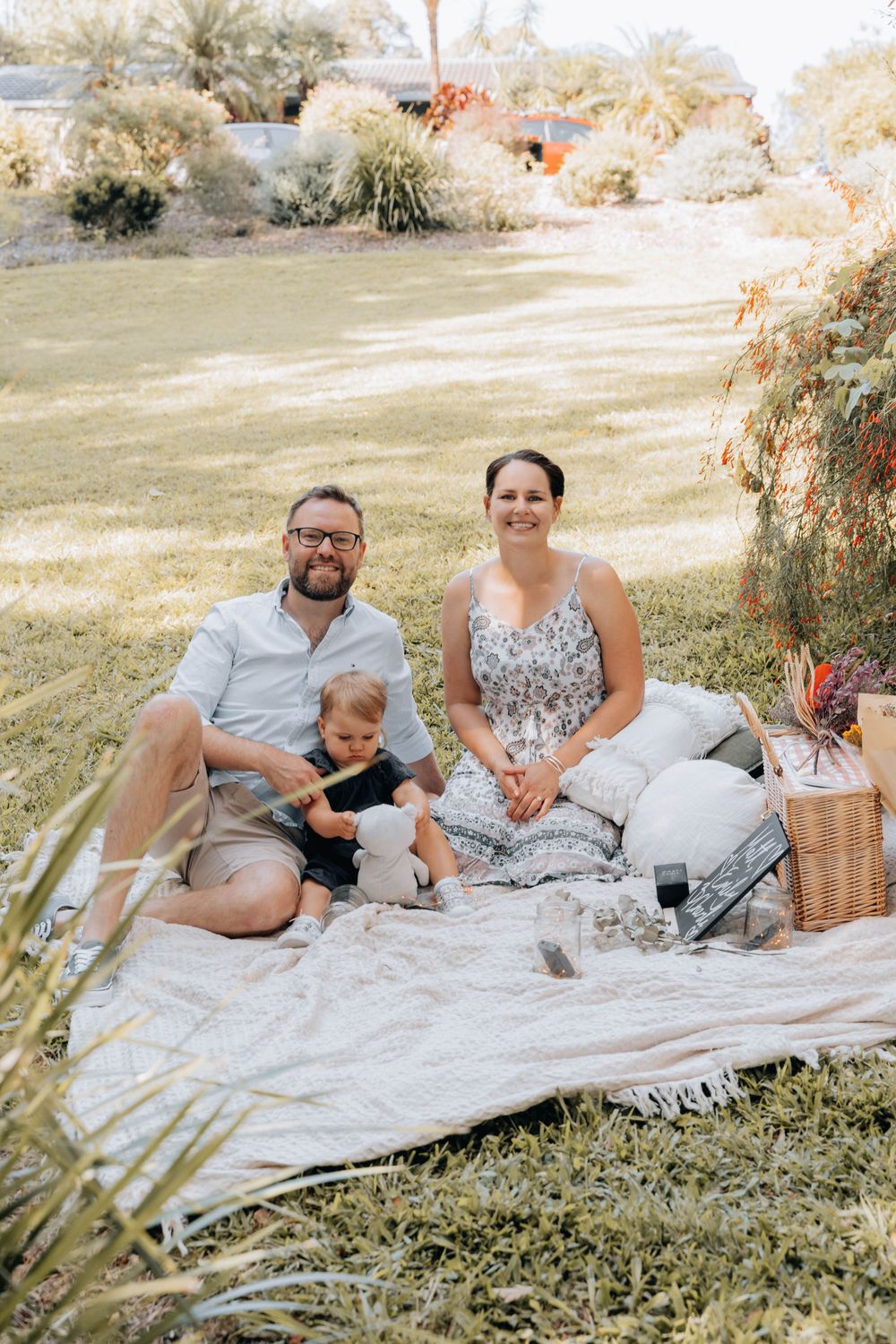 A family enjoys a sunny afternoon picnic on a blanket with a wicker basket in a grassy garden setting.