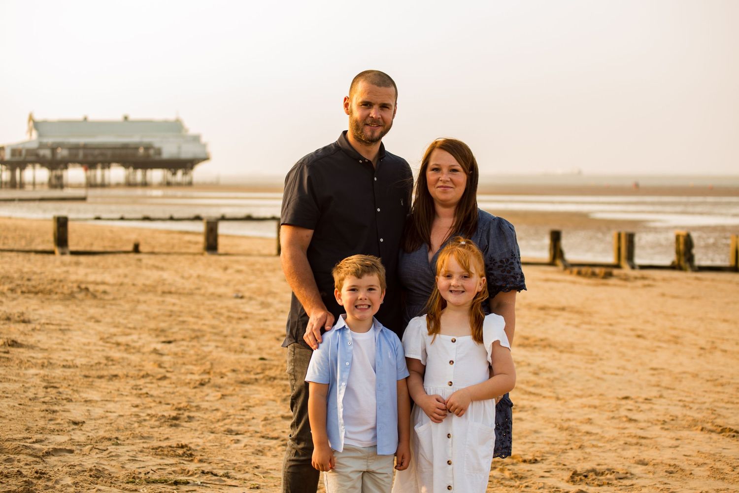 Family portrait on sandy beach with pier in background during golden hour.
