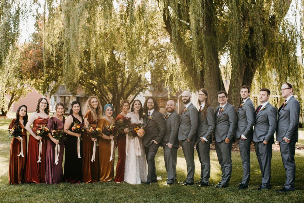 Large wedding party in autumn tones posing under weeping willow trees at outdoor fall wedding.