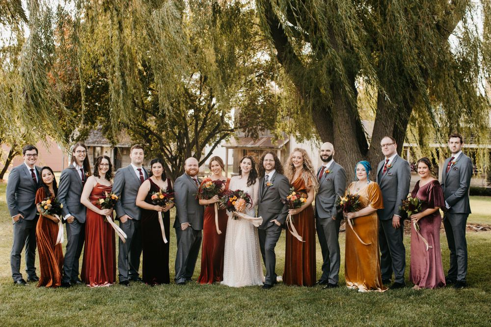 Wedding party in rust and gray attire holding fall bouquets under willow trees at autumn wedding.