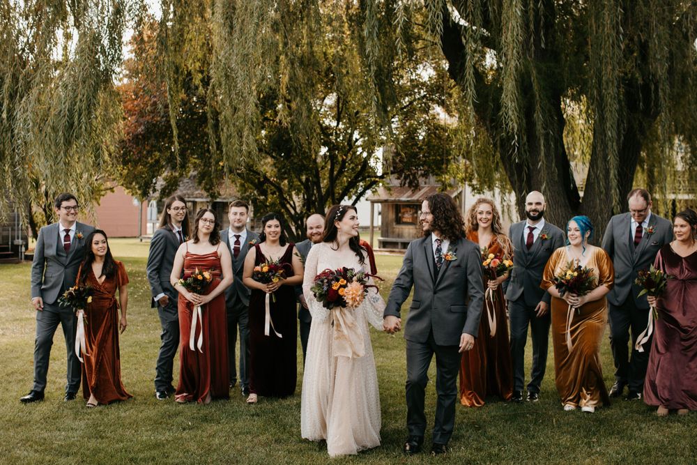 Bride and groom lead wedding party walk under weeping willows in fall outdoor wedding photos.