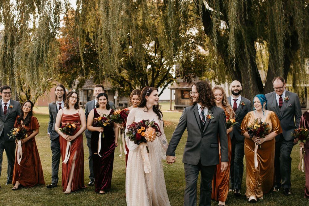 Bride and groom walk hand in hand with wedding party in fall colors under willow trees.