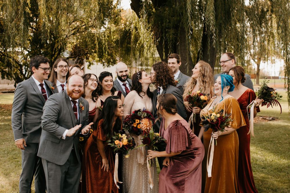 Joyful wedding party laughing together in autumn-toned attire under weeping willow trees.