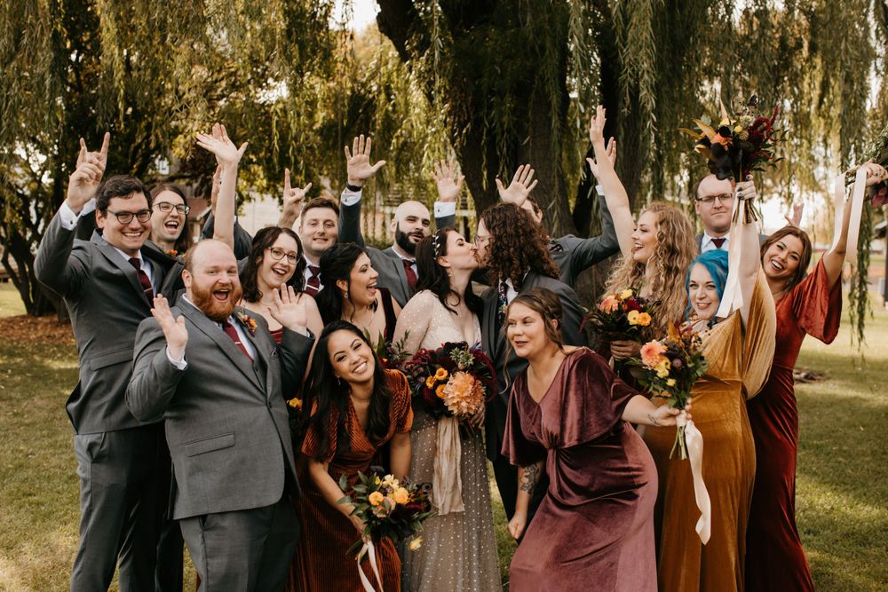 Excited wedding party cheering with hands raised in fall-colored attire under willow trees.