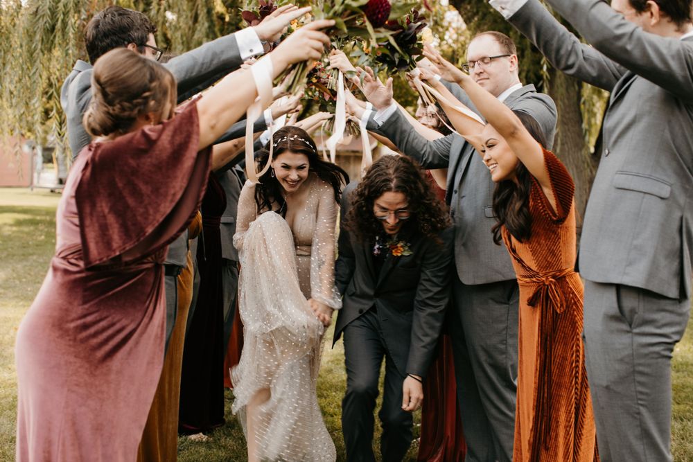 Bride and groom pass under arch of raised bouquets held by wedding party under willow tree.