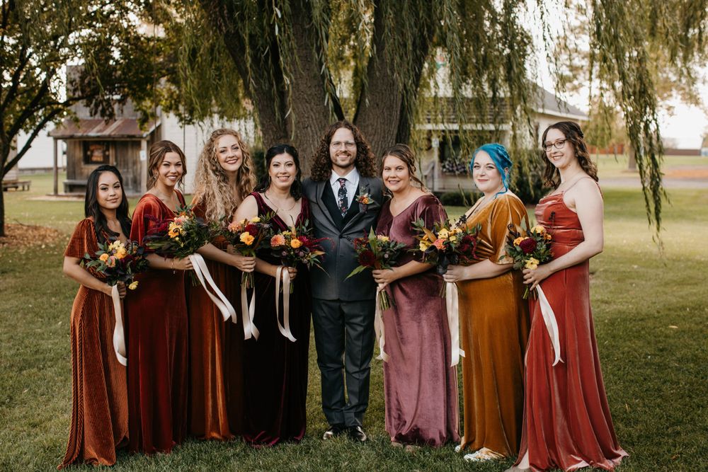 Groom poses with bridesmaids in rust and amber velvet gowns holding fall bouquets outdoors.