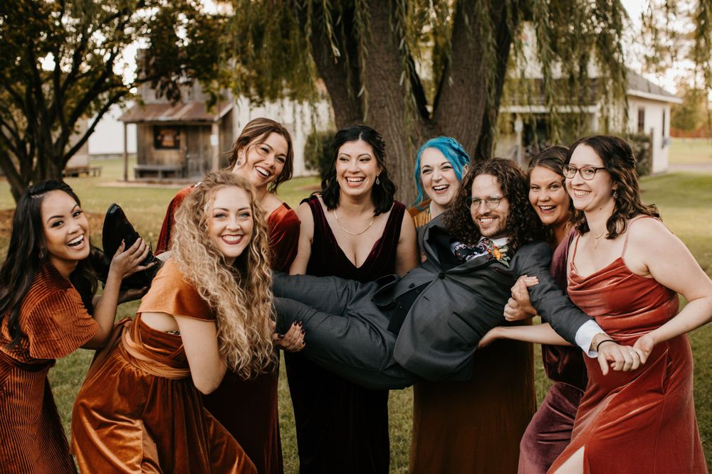 Bridesmaids in velvet rust and burgundy dresses laugh while lifting groom outdoors at fall wedding.