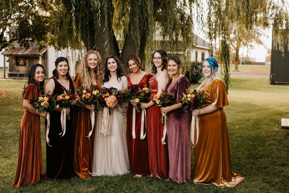 Seven bridesmaids in mismatched velvet fall-toned dresses hold bouquets under willow tree.