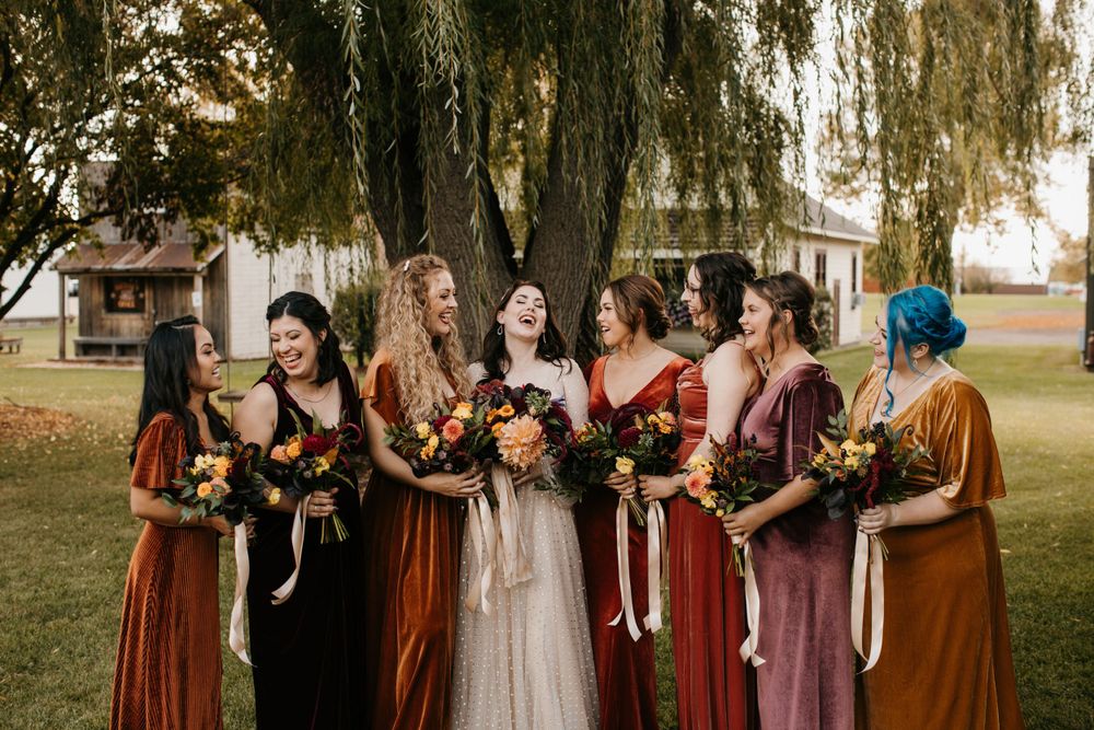 Bridesmaids in velvet autumn-colored gowns laugh together holding floral bouquets by willow tree.