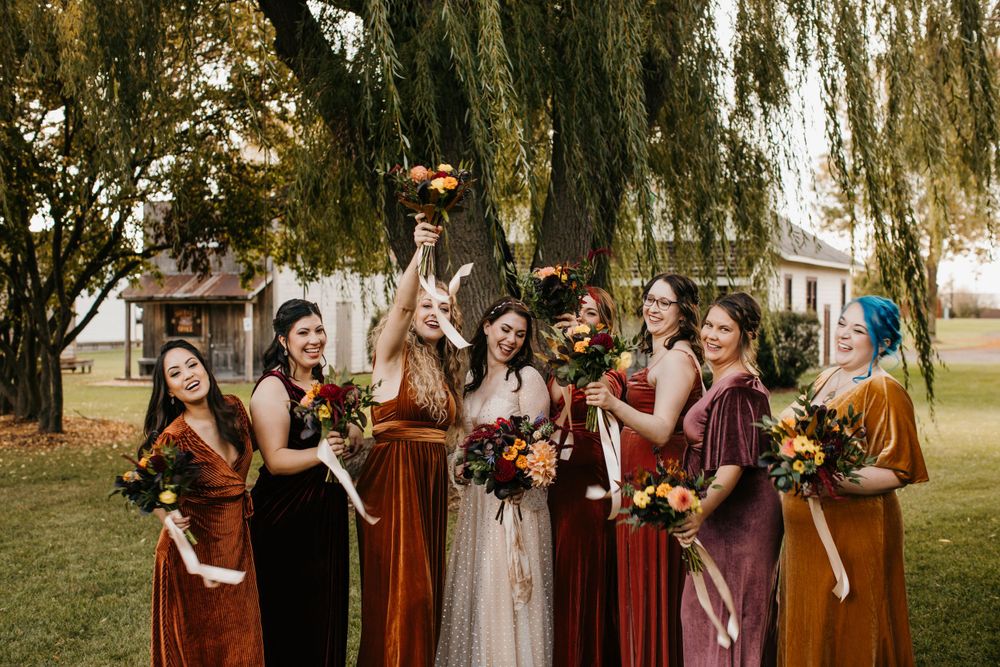 Bride and bridesmaids in rust velvet dresses celebrate raising bouquets under willow tree.